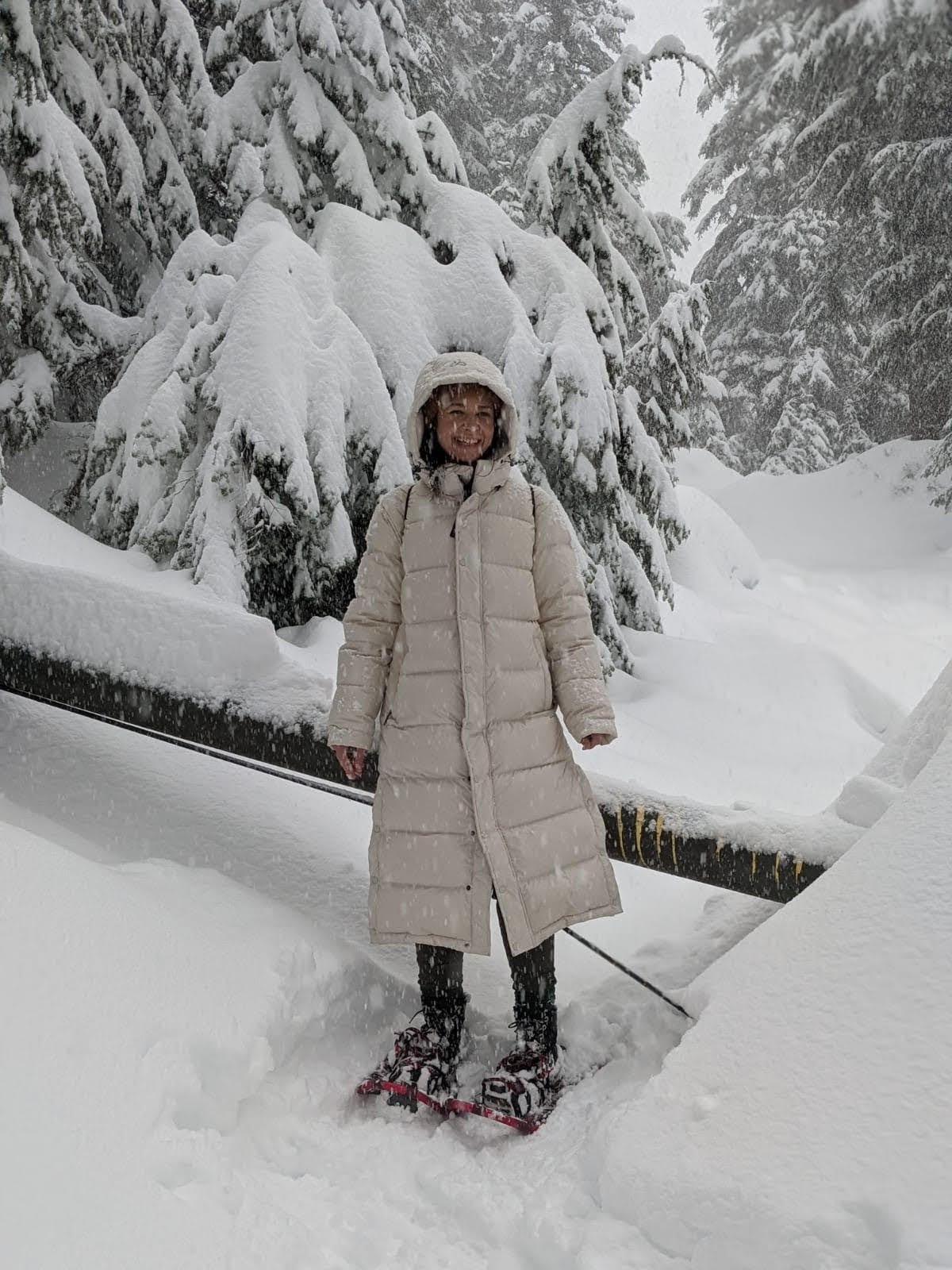 default-34 A woman in a white poofy jacket and snowshoes smiles as she poses on a snow-covered trail through tall pine trees.