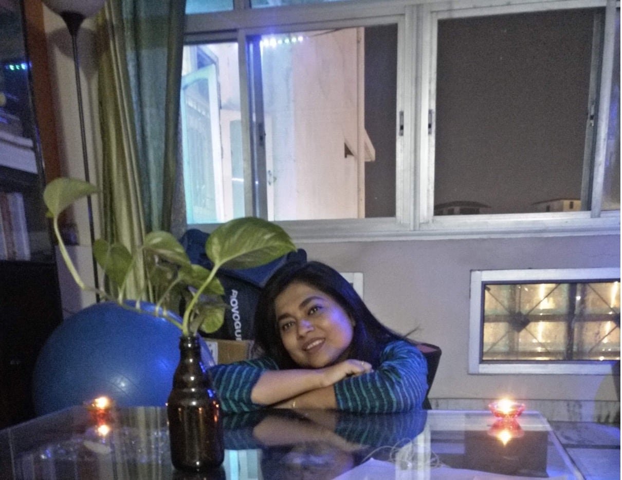 Large windows can be seen in the background as a woman sits, her arms resting on a clear kitchen table.