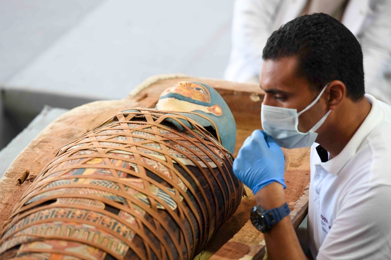 default-539 An archaeologist inspects an Egyptian mummy that is wrapped in an elaborate and colourful burial shroud. The archaeologist is wearing a medical mask and gloves.