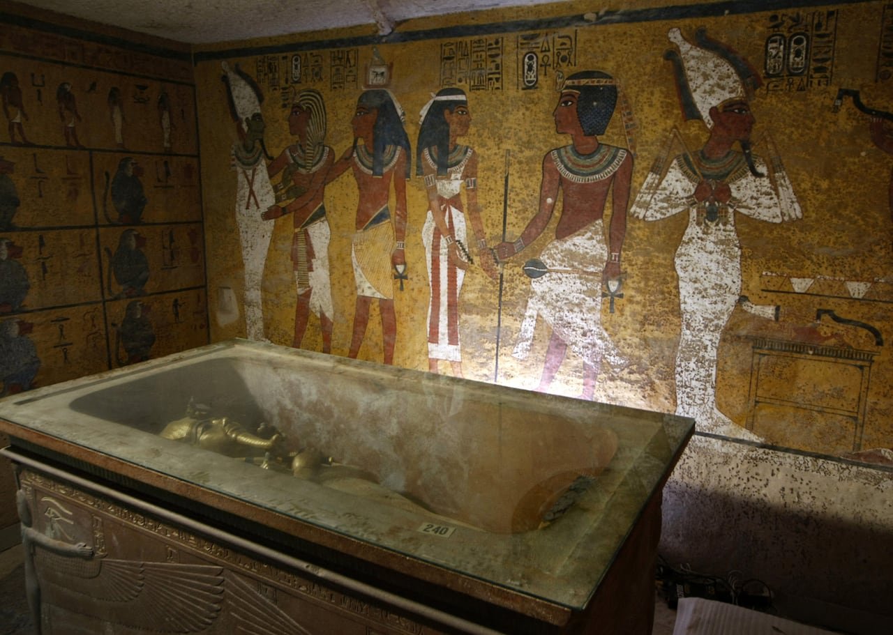 default-540 A golden sarcophagus is shown lying in an elaborate burial chamber. The wall behind it is colourfully painted with figures.
