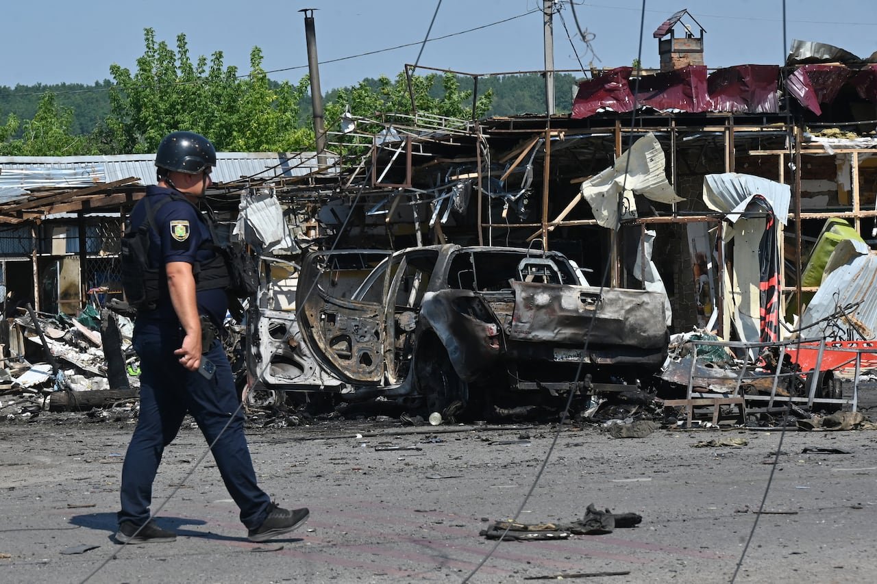 A police officer is shown walking in an area filled with debris and damaged vehicles and structures.