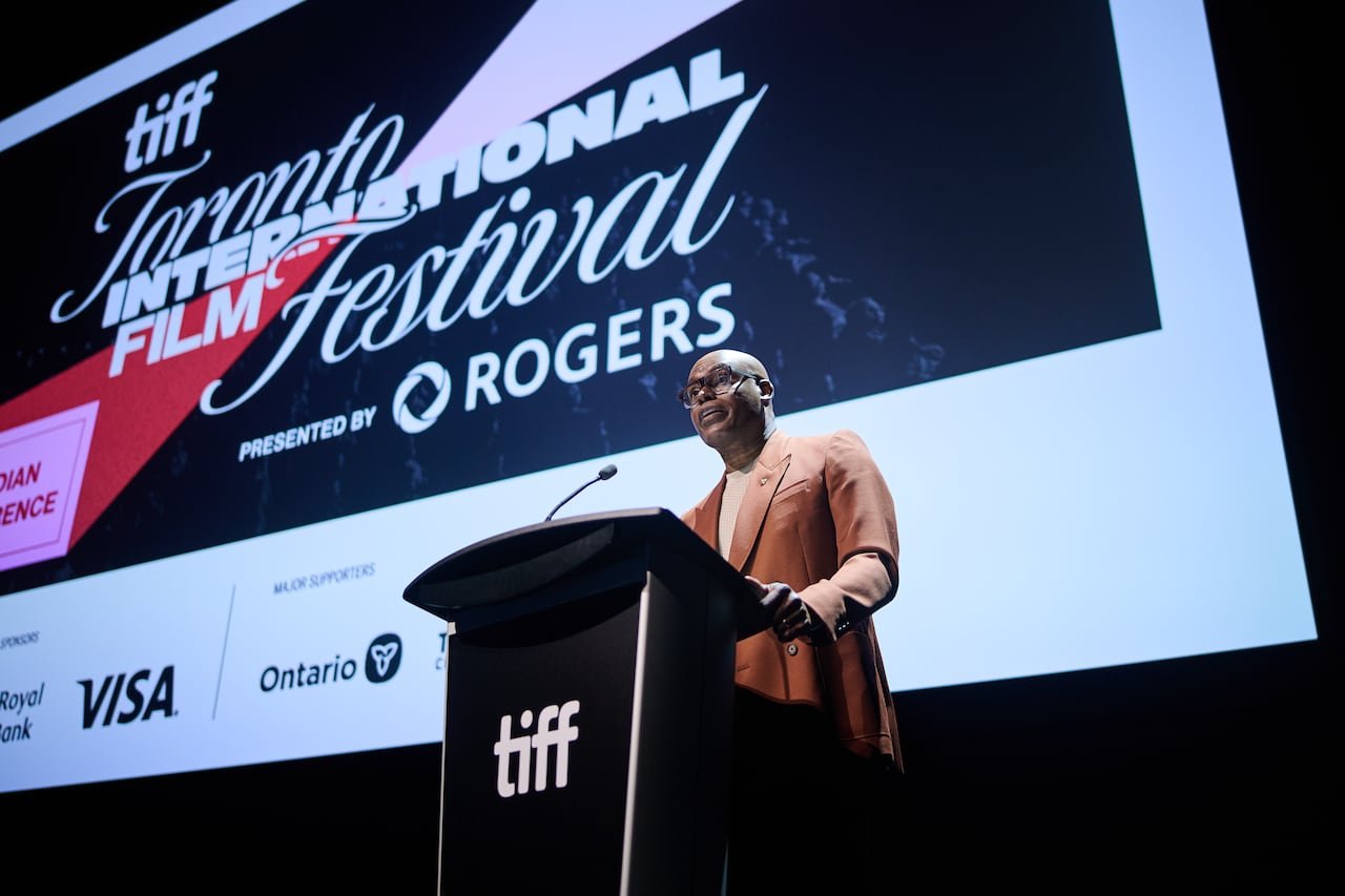 default-886 A bald man wearing glasses speaks at a podium in front of a large screen advertising the Toronto International Film Festival.