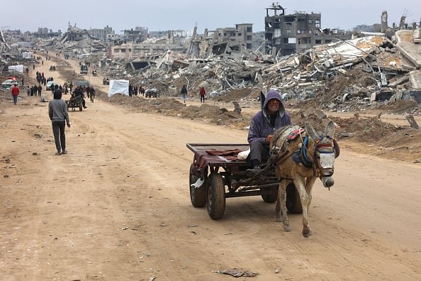 A man rides in a donkey-pulled cart past the rubble of destroyed buildings.